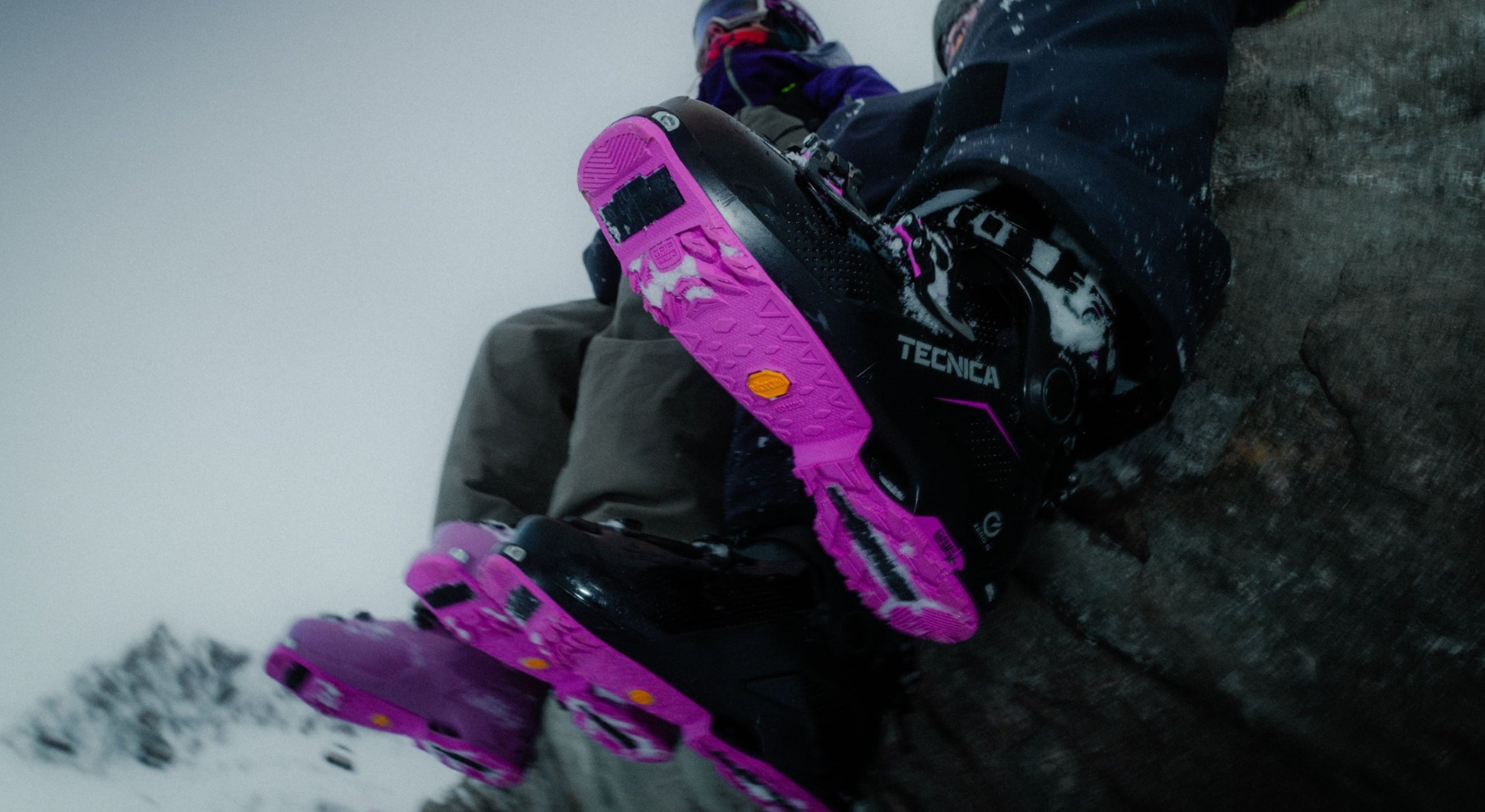 Close-up of a person sitting on a snowy mountain rock, wearing black and white Tecnica ski boots with bright pink snowboard bindings and bindings attached to the ski edges.
