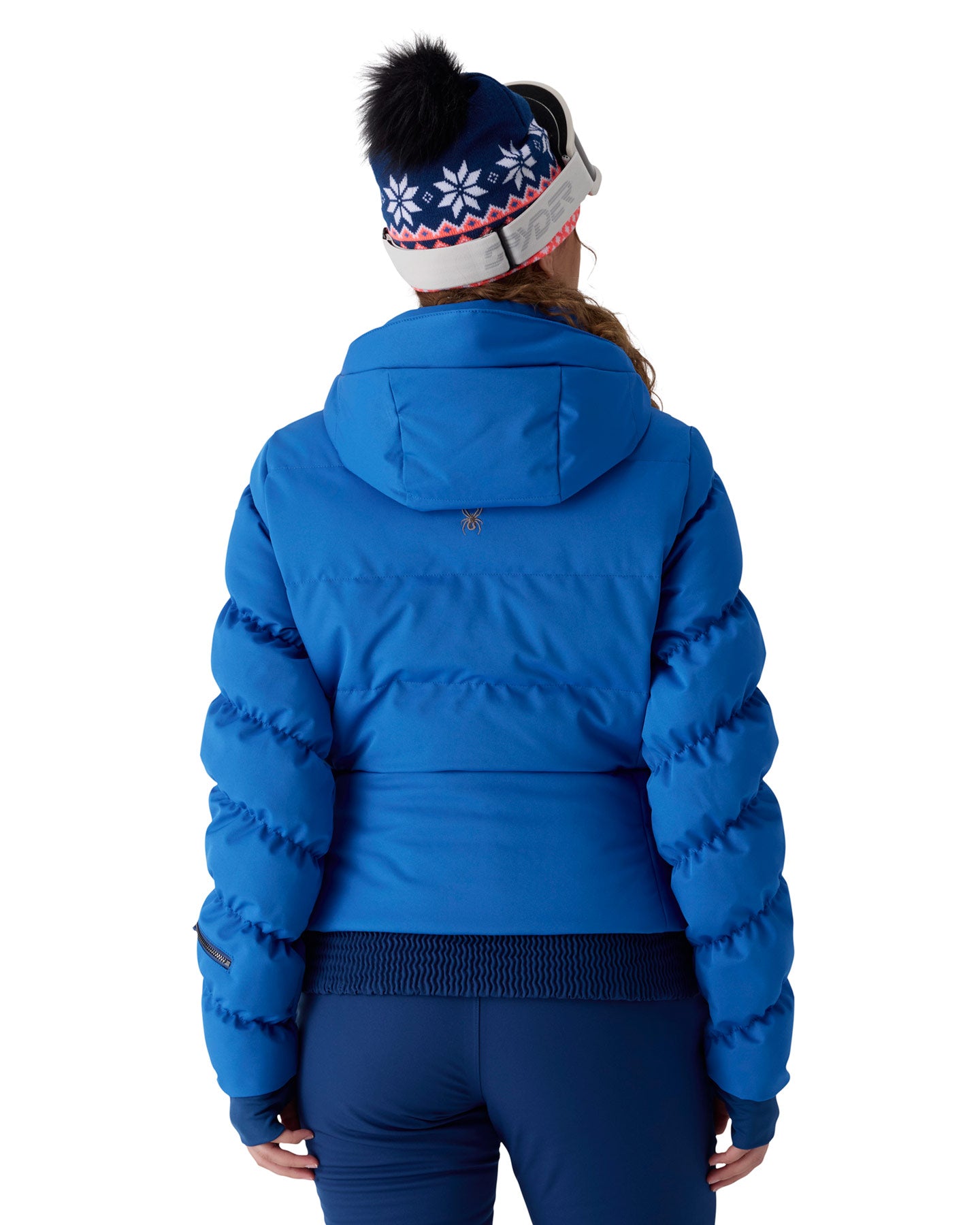 A woman viewed from behind wearing a bright blue puffer jacket, dark blue leggings, and a navy knit beanie with white snowflakes and a black pom-pom, against a light gray studio background.