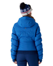 A woman viewed from behind wearing a bright blue puffer jacket, dark blue leggings, and a navy knit beanie with white snowflakes and a black pom-pom, against a light gray studio background.