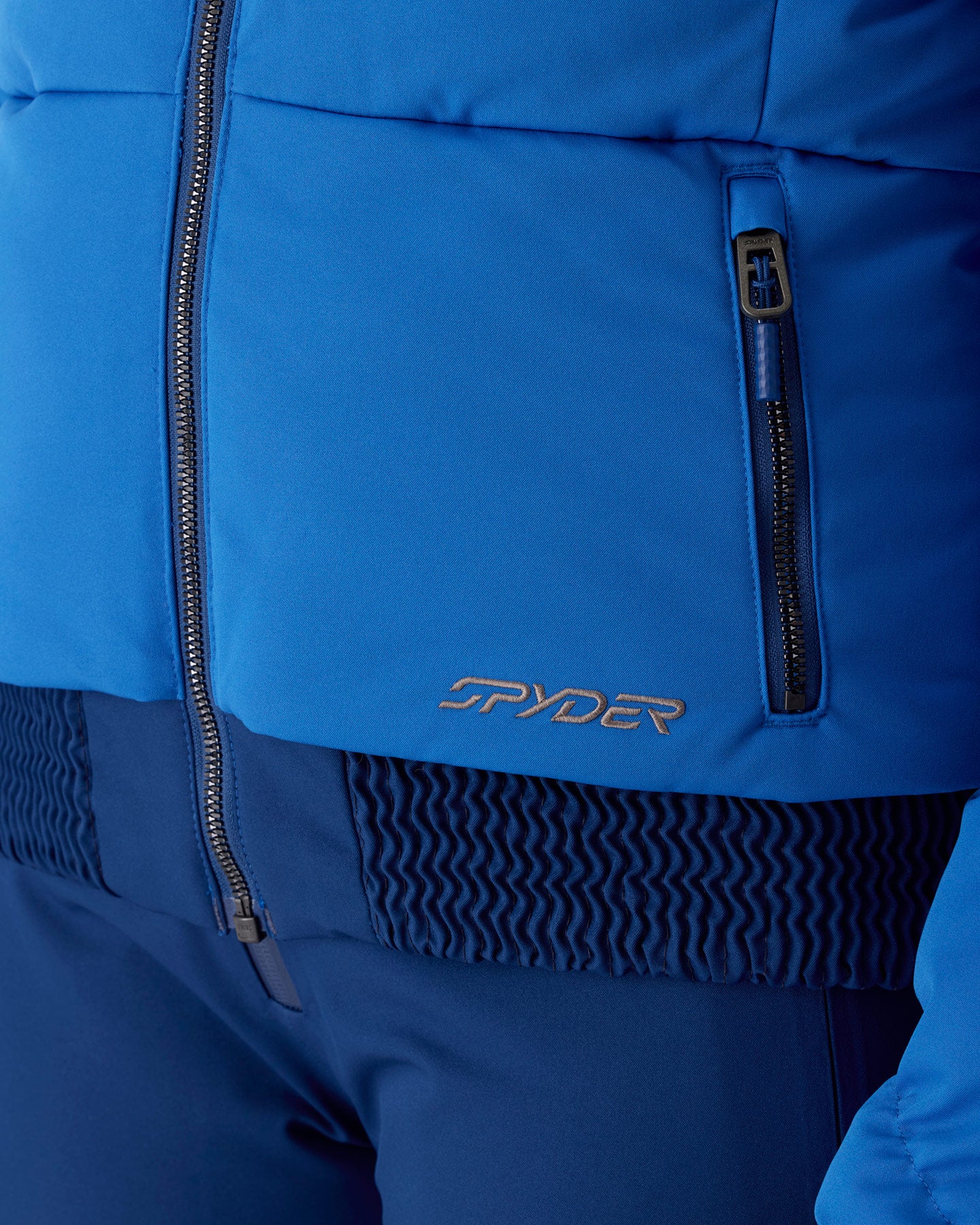 Close-up of a blue ski jacket showing the front zipper, a vertical zip pocket, and the Spyder logo embroidered in metallic gold on the lower front, paired with matching blue ski pants.