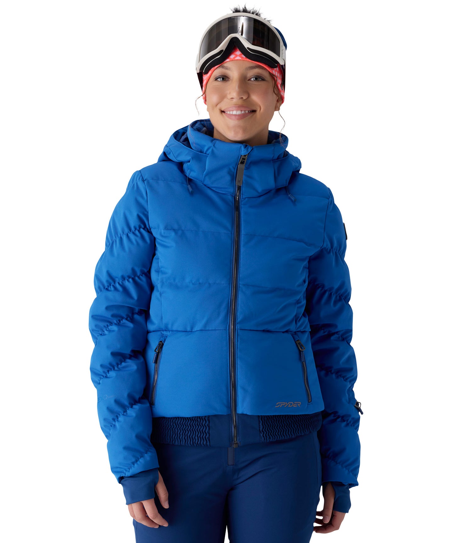 A woman wearing a bright blue hooded puffer jacket, blue ski pants, ski goggles, and a red bandana, photographed against a plain white studio background.