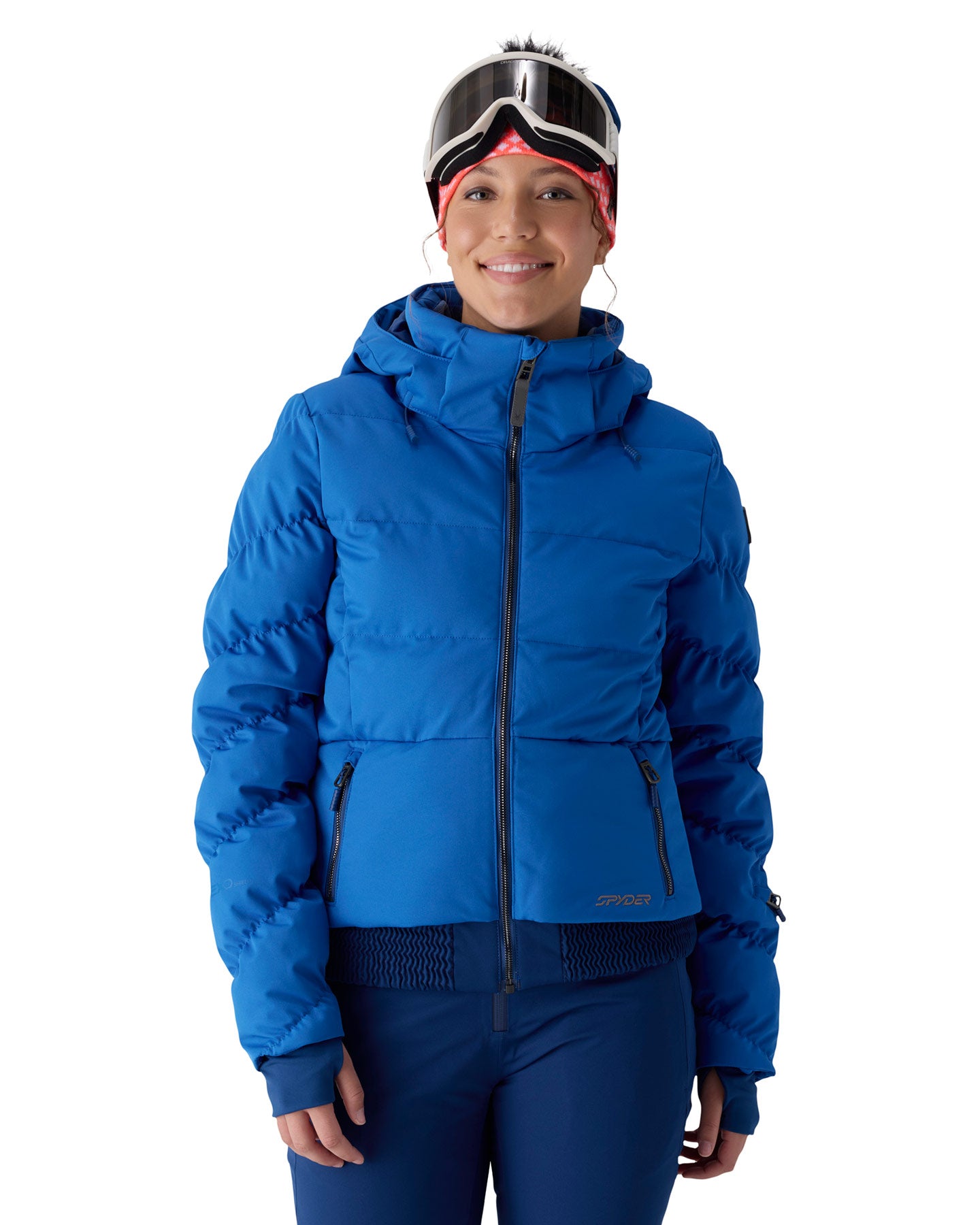 A woman wearing a bright blue hooded puffer jacket, blue ski pants, ski goggles, and a red bandana, photographed against a plain white studio background.