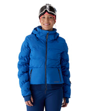 A woman wearing a bright blue hooded puffer jacket, blue ski pants, ski goggles, and a red bandana, photographed against a plain white studio background.